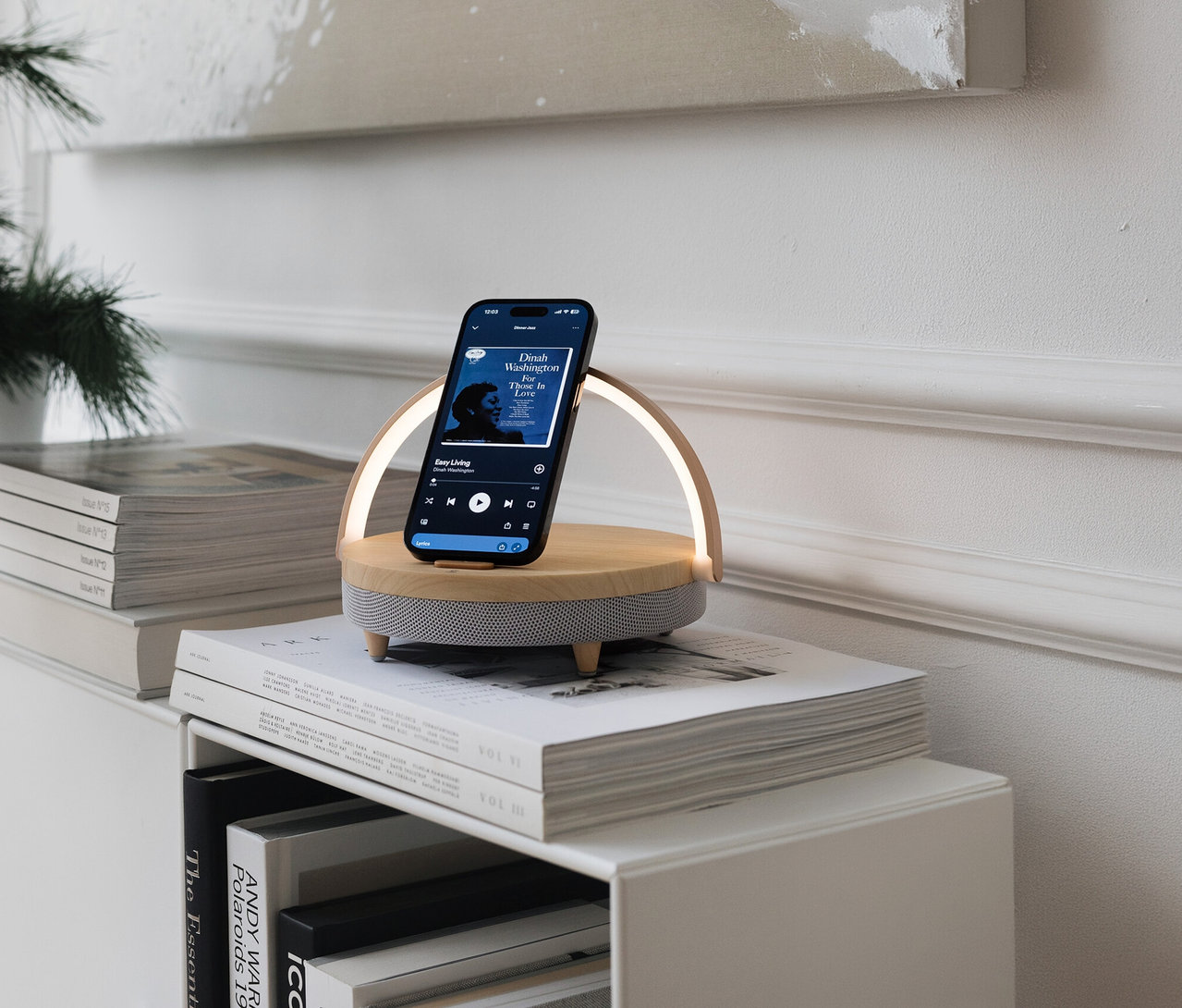Handy auf Bluetooth-Lautsprecher mit kabellosem Ladegerät und LED-Nachtlicht auf Bücherstapel. Handy zeigt Musik-App.