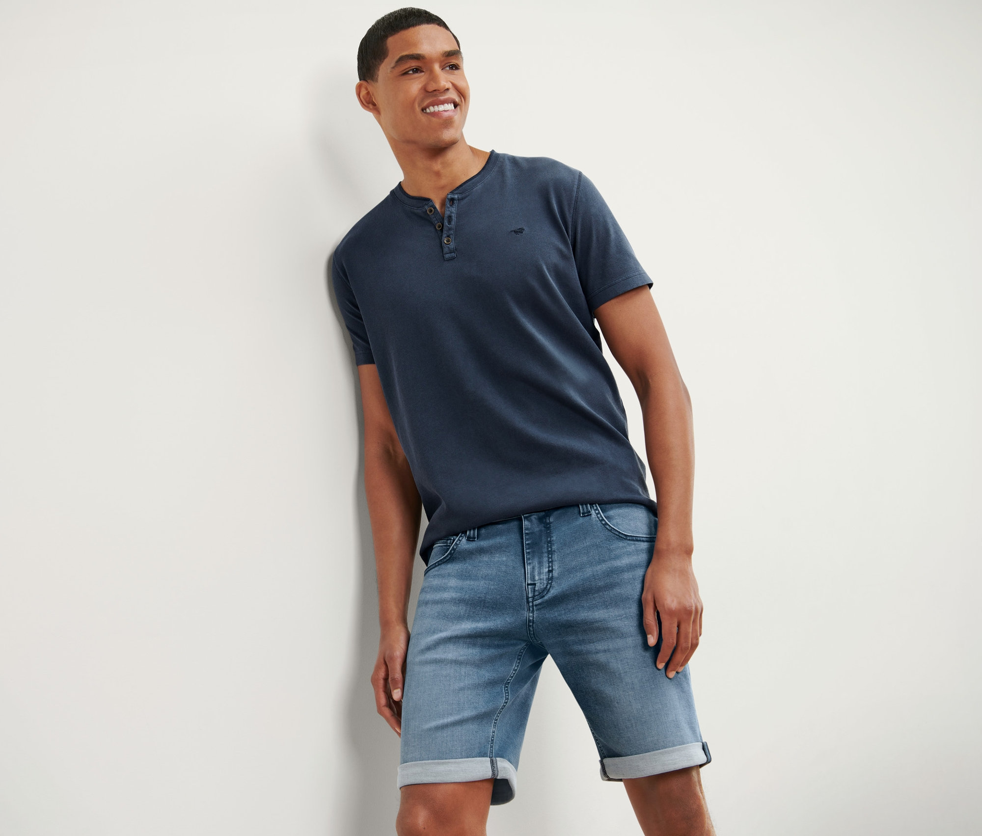 Ein lächelnder Mann lehnt an einer Wand und trägt ein blaues T-Shirt und Jeans-Shorts »Mustang«.