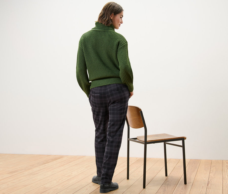 Eine Frau steht mit dem Rücken zur Kamera und trägt einen grünen Pullover und eine karierte Joggpants.