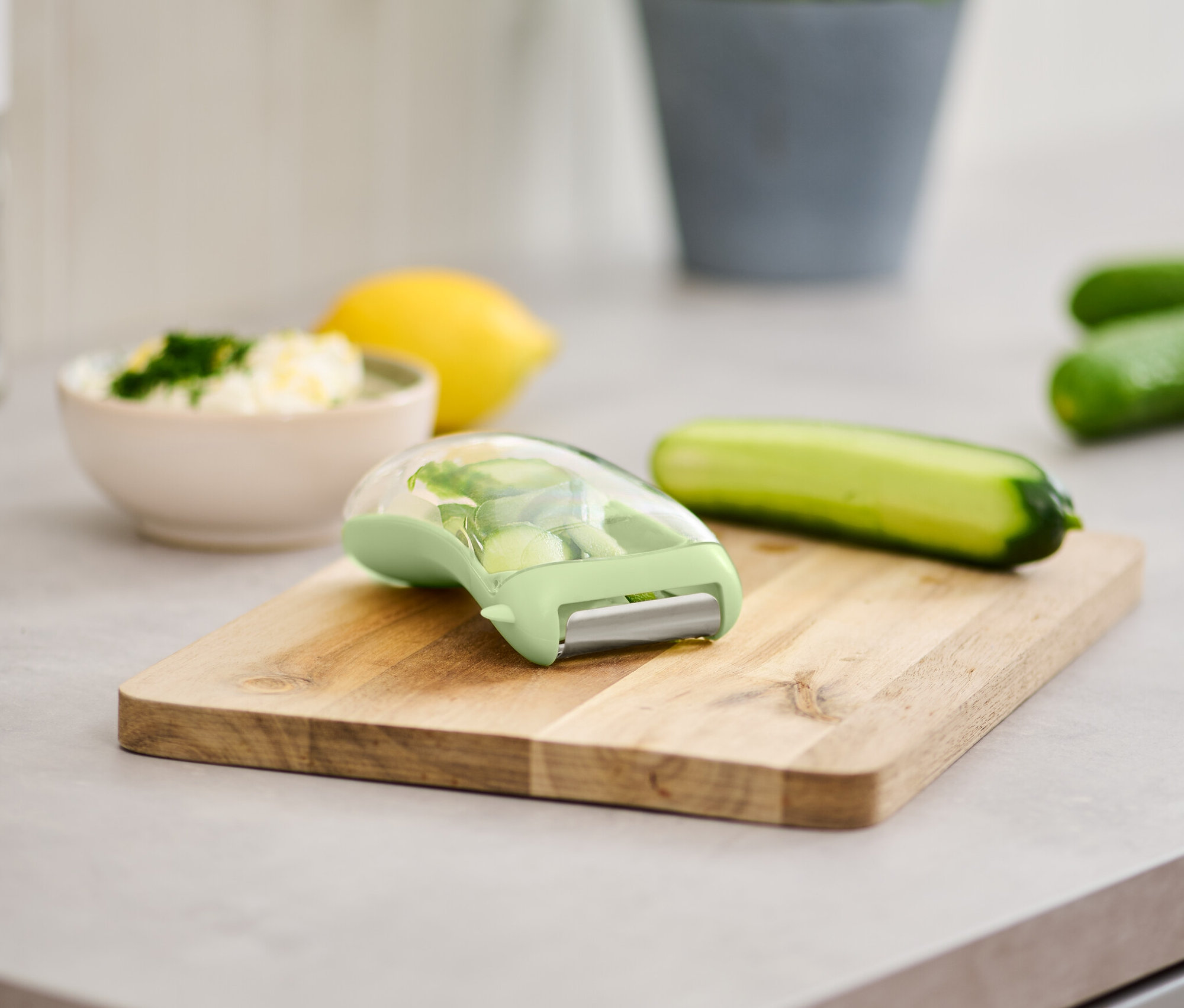 Universalschäler mit Auffangbehälter auf einem Holzbrett neben Gurke und einer Schüssel mit Essen.