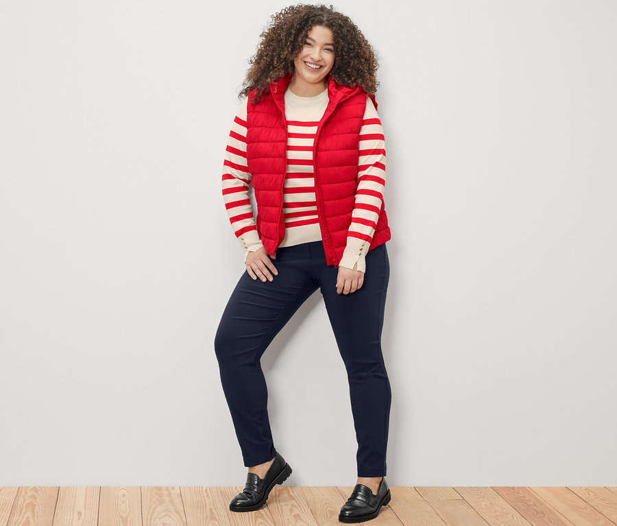 Eine Frau steht und lächelt in die Kamera, sie trägt einen rot gestreiften Feinstrickpullover, eine rote Steppweste mit Kapuze und eine dunkelblaue Bengalin-Hose.