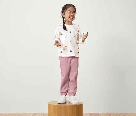 Ein Mädchen mit Zopf steht auf einem Holzsockel. Sie trägt ein weißes Langarmshirt mit bunten Herzen und eine rosafarbene Hose.