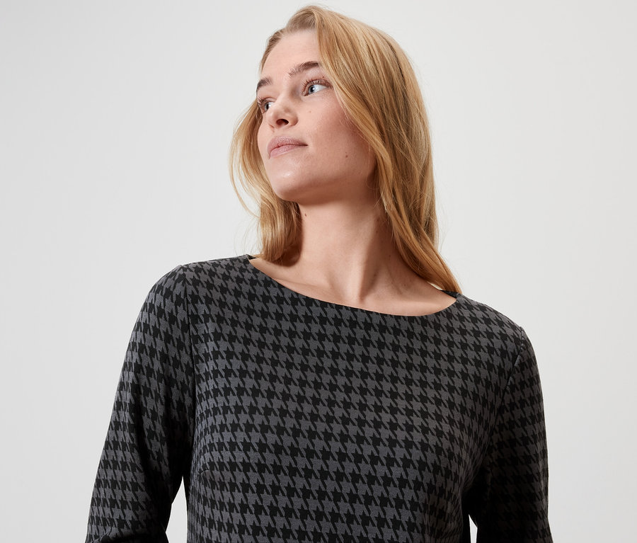 Blonde Frau mit langen Haaren trägt ein Kleid mit Hahnentrittmuster.