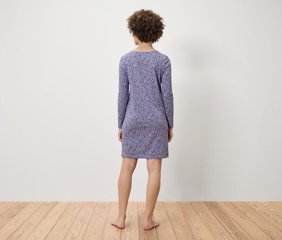 Eine Frau mit lockigem Haar steht mit dem Rücken zur Kamera und trägt ein blaues, langärmliges Kleid mit Blattmuster.