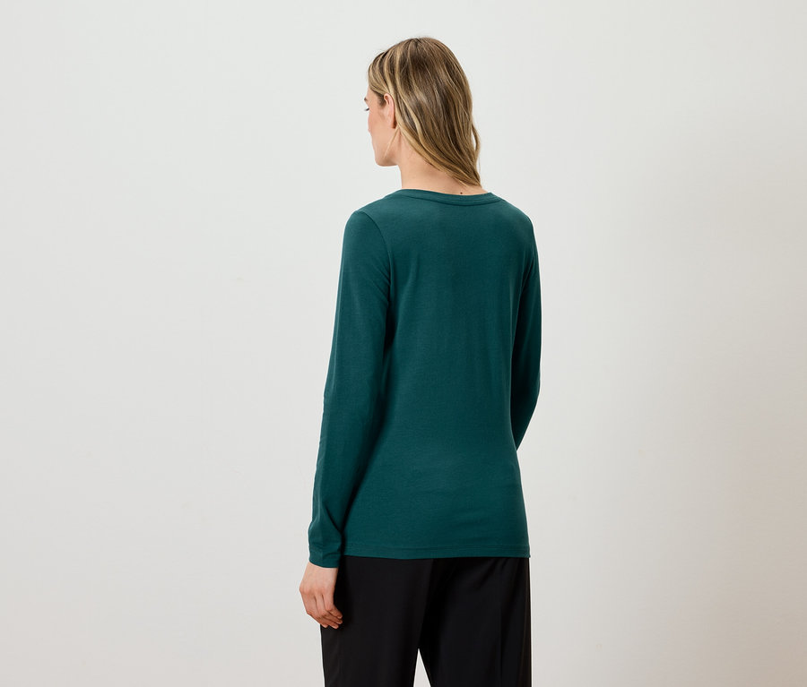 Von hinten ist eine Frau mit blonden Haaren zu sehen, die ein grünes Langarmshirt und eine schwarze Hose trägt.