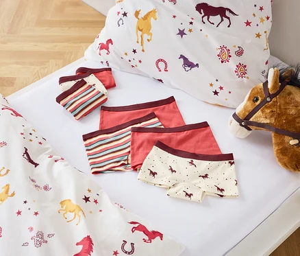 Auf einem weißen Bett liegen Bettwäsche mit Pferdeaufdruck, ein Set Kinderunterwäsche und ein Steckenpferd.