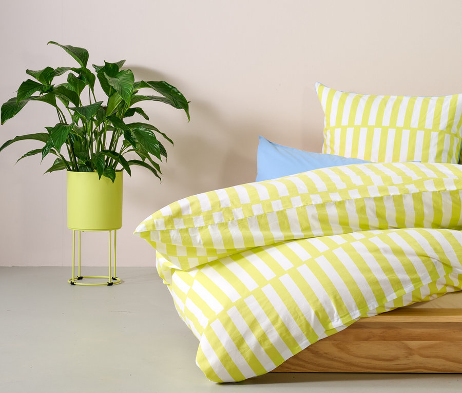 Grüne Pflanze in runder Pflanzbox steht neben Bett mit gelb gestreifter Bettwäsche.
