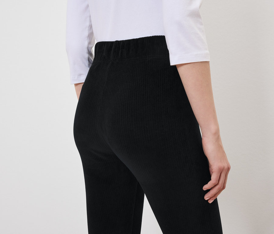 Von hinten ist eine Frau in einer schwarzen Cordhose und einem weißen T-Shirt zu sehen.