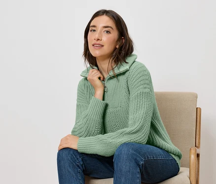 Eine Frau sitzt auf einem Stuhl und trägt einen grünen Pullover und blaue Jeans.