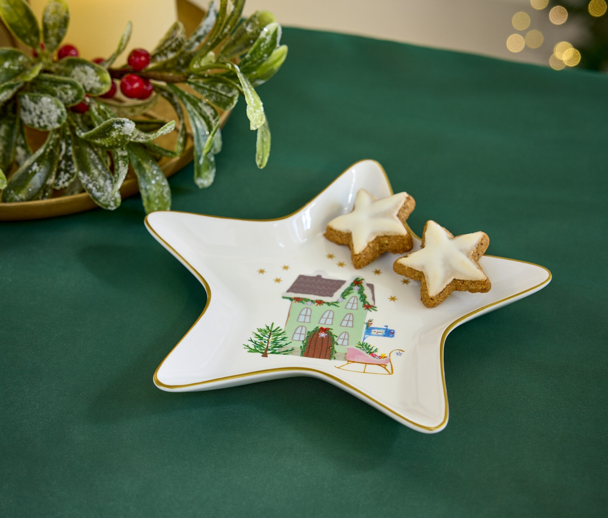 Auf einer grünen Tischdecke liegt ein sternförmiger Teller mit einem Weihnachtsmotiv und zwei sternförmige Kekse. Im Hintergrund sind eine Kerze und Weihnachtsdekoration zu sehen.