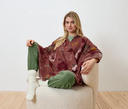 Eine blonde Frau sitzt in einem Sessel und trägt einen geblümten Kimono, eine grüne Hose und weiße Socken.