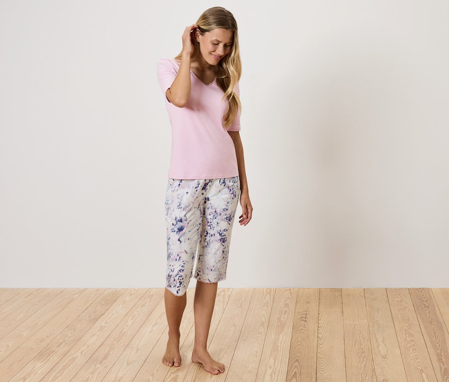 Frau mit rosa T-Shirt und geblümter Hose steht auf Holzboden.