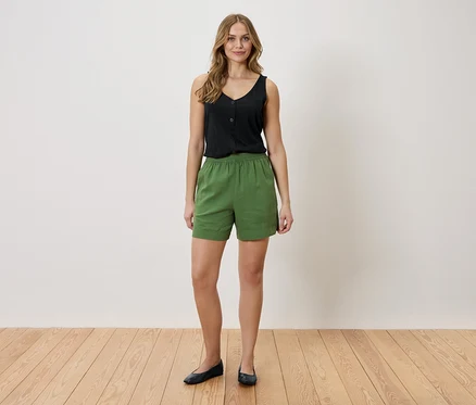 Eine Frau steht vor einer weißen Wand. Sie trägt ein schwarzes Top und grüne Shorts.