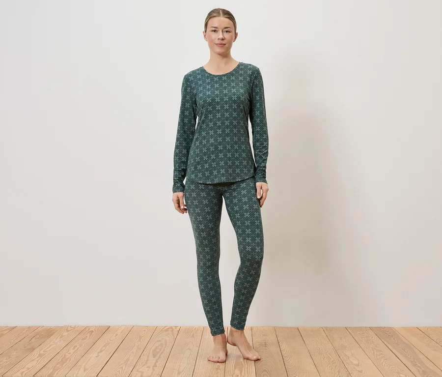 Eine Frau steht barfuß vor einer weißen Wand auf einem Holzboden und trägt einen dunkelgrünen Pyjama mit einem hellgrünen Muster.