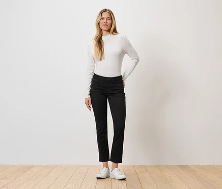 Eine Frau steht vor einer Wand, sie trägt ein langärmliges Oberteil, schwarze Jeans und weiße Turnschuhe.