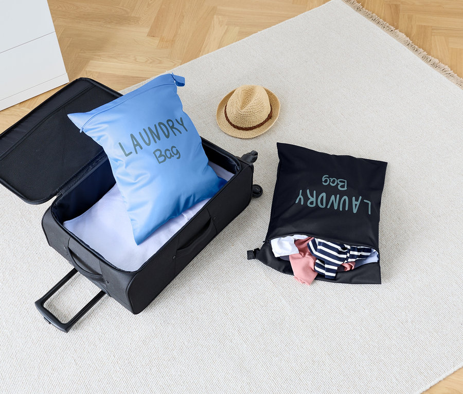 Auf einem Teppich steht ein offener Koffer mit einem blauen Wäschesack darin, neben einem schwarzen Wäschesack und einem Hut.