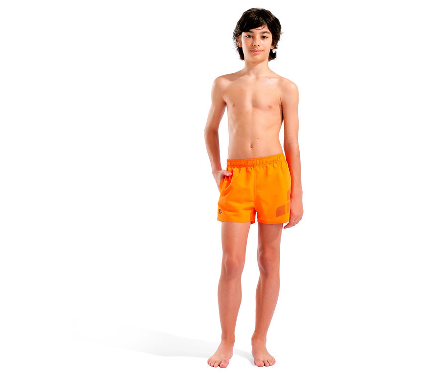 Ein barfüßiger Junge steht vor einem weißen Hintergrund und trägt orangefarbene Badehosen.
