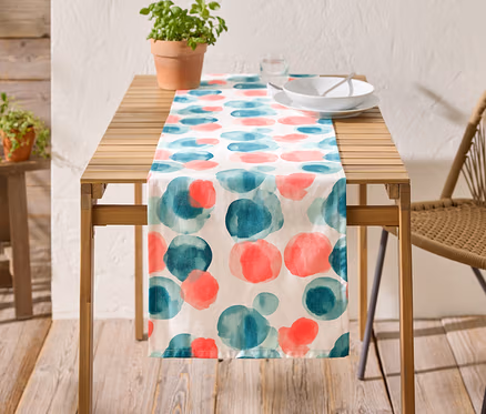 Ein Holztisch mit einer Tischdecke mit blauen und roten Kreisen, Geschirr und einer Topfpflanze darauf.