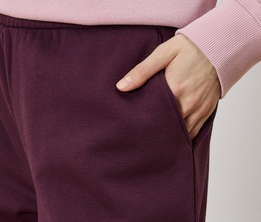 Nahaufnahme einer Person, die weinrote Trainingshose und einen rosa Pullover trägt und ihre Hand in die Hosentasche steckt.