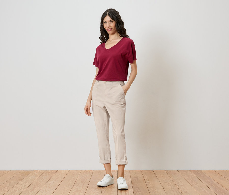 Eine Frau steht vor einer Wand und trägt ein rotes T-Shirt, helle Hosen und weiße Turnschuhe.