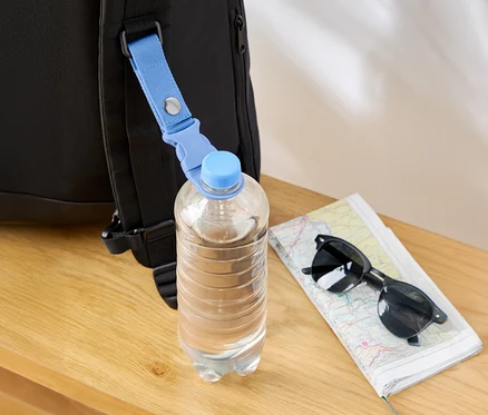 Eine Wasserflasche hängt mit einem blauen Band an einem schwarzen Rucksack, daneben liegen eine Karte und eine Sonnenbrille.