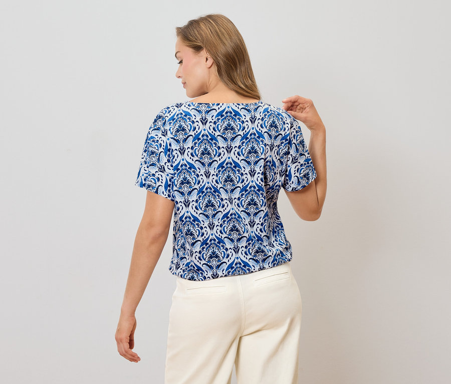 Rückansicht einer Frau mit blonden Haaren, die eine blau-weiß gemusterte Bluse und eine helle Hose trägt.