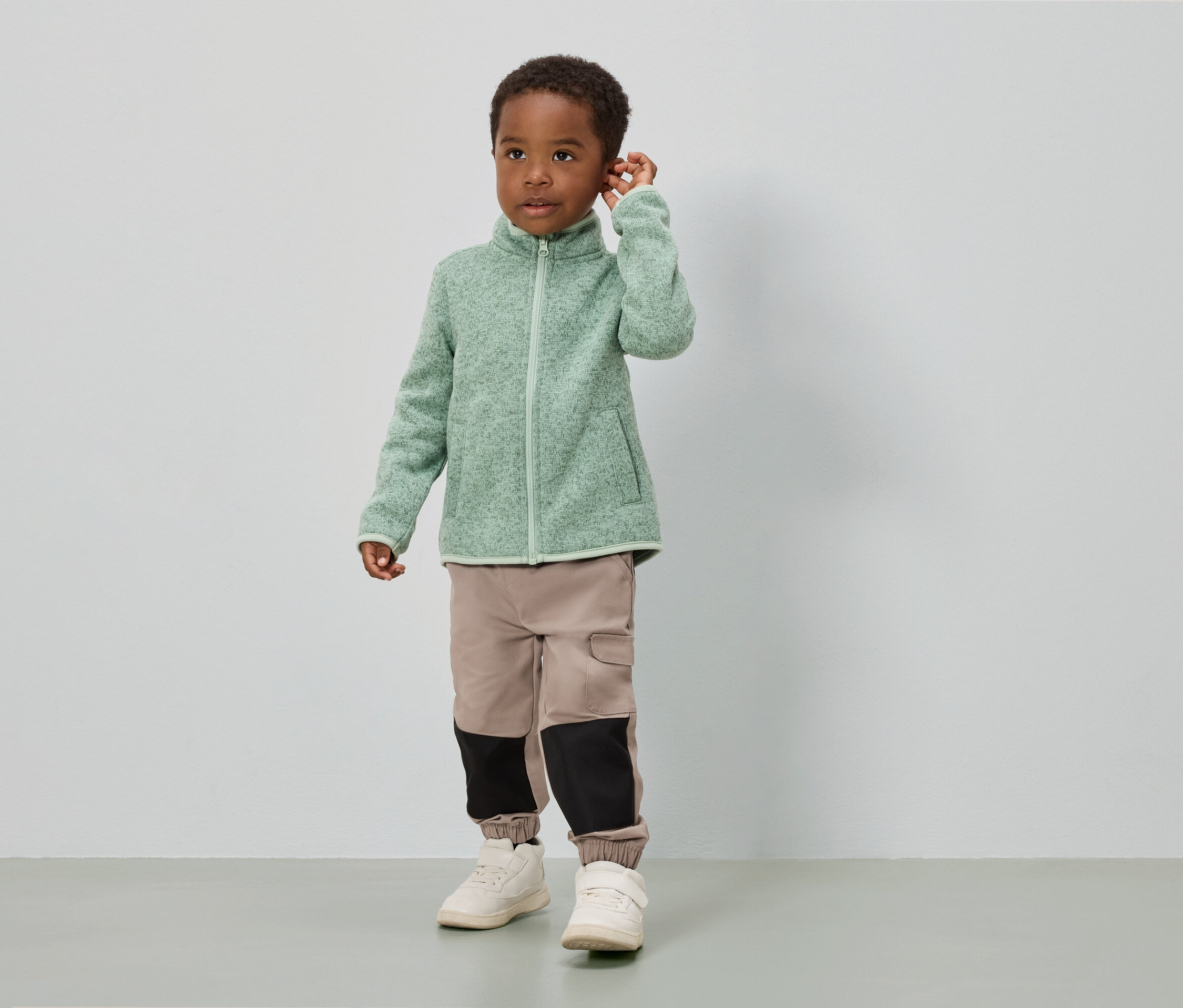 Ein Junge steht vor einer weißen Wand und trägt eine hellgrüne Fleecejacke, beige Hosen mit schwarzen Einsätzen und weiße Schuhe.