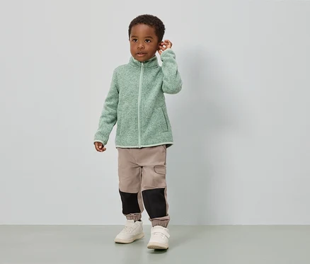 Ein Junge steht vor einer weißen Wand und trägt eine hellgrüne Fleecejacke, beige Hosen mit schwarzen Einsätzen und weiße Schuhe.