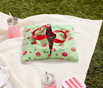 Picknickdecke mit Erdbeermuster-Tasche gefüllt mit Beeren, Getränken und Besteck auf Gras.