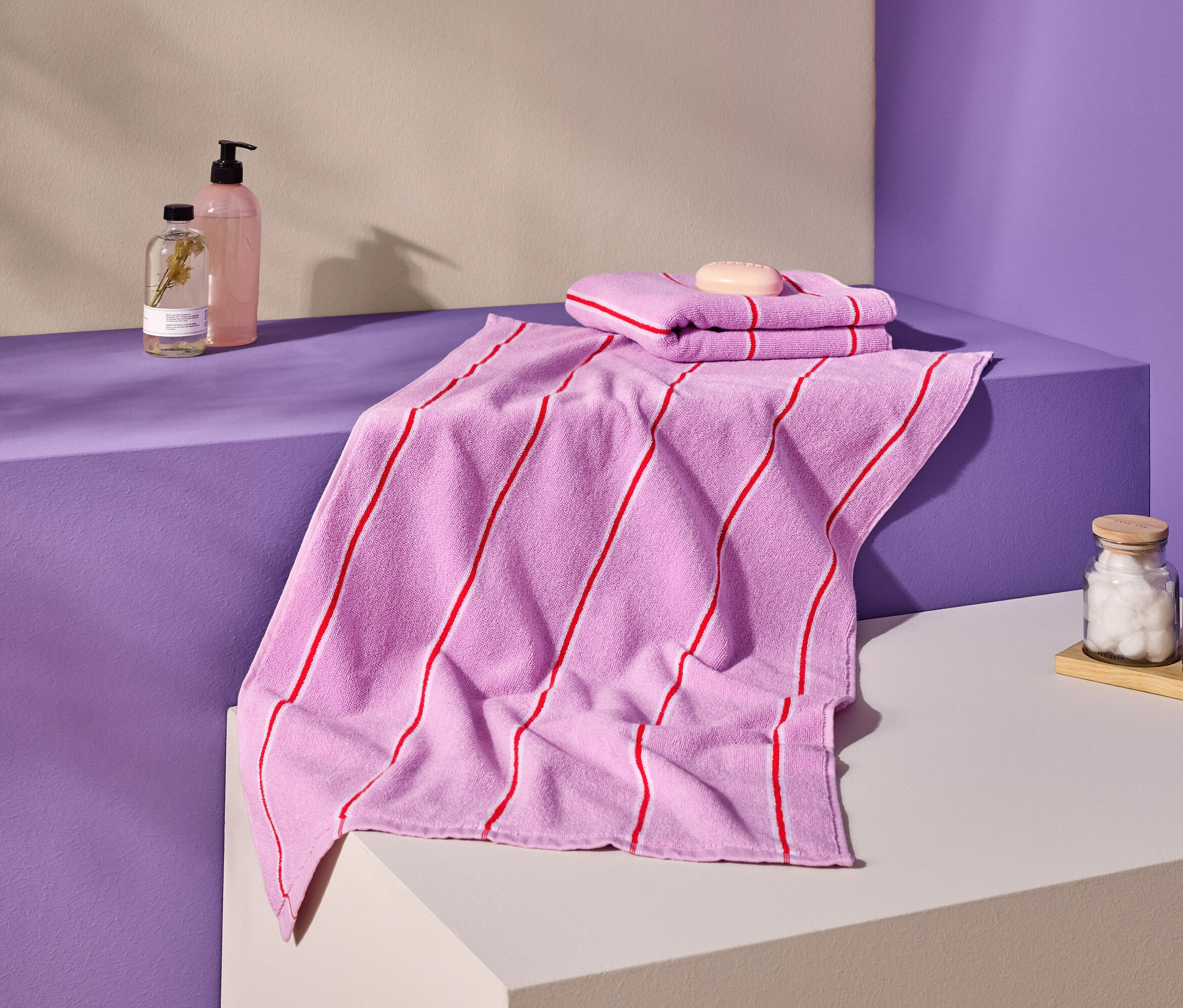 Aufnahme von rosa Handtüchern mit roten Streifen, Seife, Flaschen und Behältern auf einer lila und cremefarbenen Oberfläche.