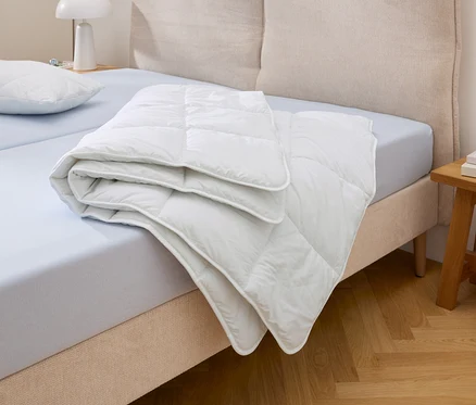 Ein weißes Steppbett Cell Solution® in Normalgröße liegt gefaltet auf einem hellblauen Bett neben einem Kissen.