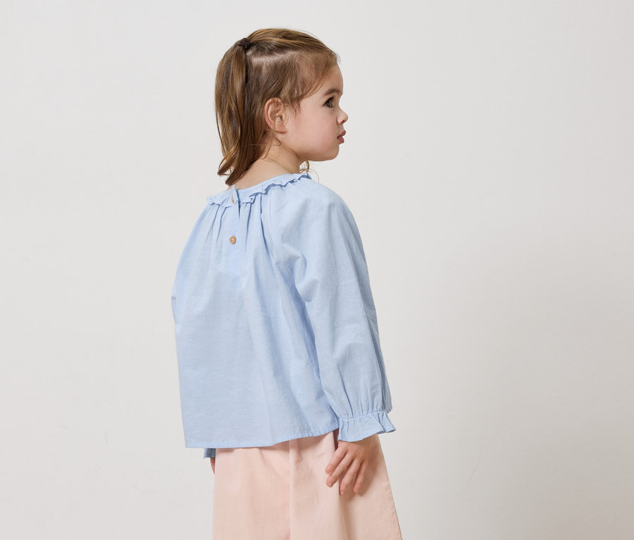 Mädchen von hinten seitlich mit blauem Rüschenhemd und rosafarbener Hose vor weißem Hintergrund.