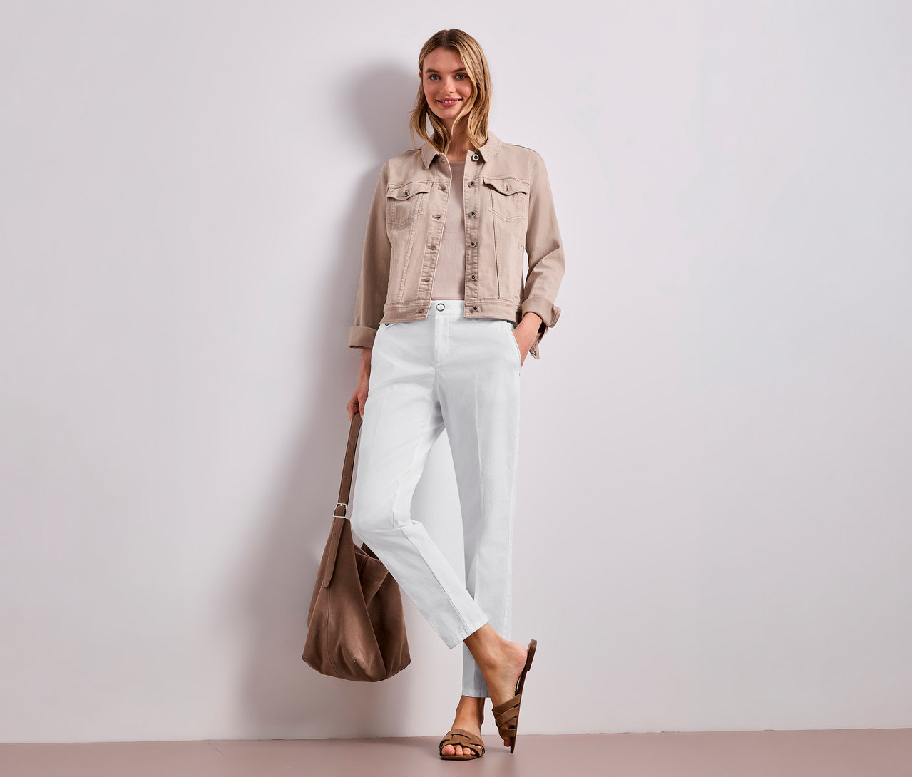 Eine lächelnde Frau steht vor einer weißen Wand und trägt eine beigefarbene Jeansjacke, ein helles Oberteil und eine weiße Street One Chino Hose. Sie hält eine braune Umhängetasche und trägt braune Sandalen.