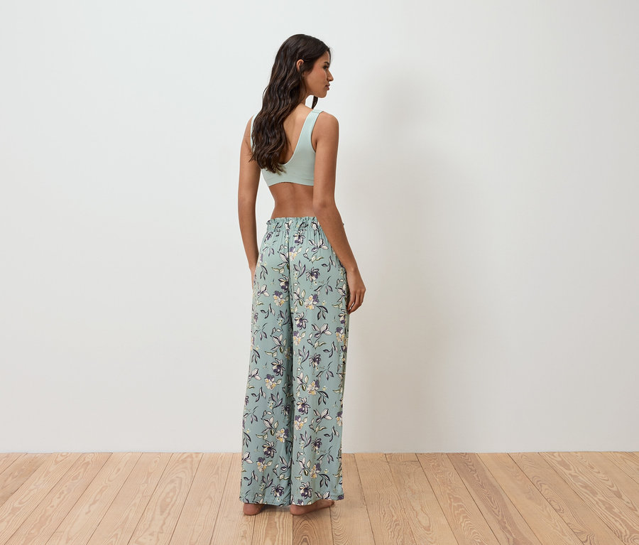 Eine Frau steht mit dem Rücken zur Kamera und trägt ein hellgrünes Top und eine locker sitzende, blumengemusterte Hose.