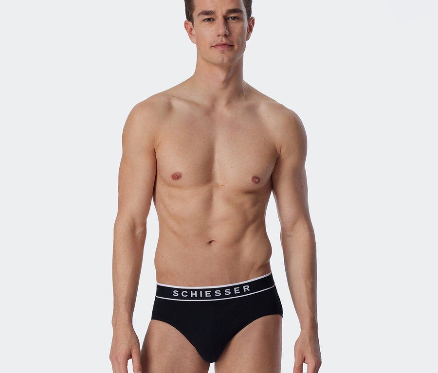 Oberkörperfreier Mann trägt schwarze SCHIESSER Rio-Slips mit weißem Logo-Bund.