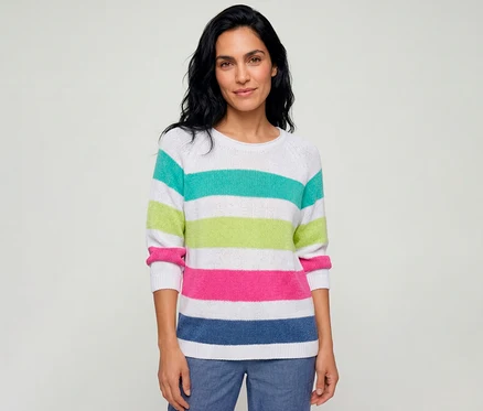 Eine Frau trägt einen gestreiften Olsen Pullover in Weiß mit türkisen, lindgrünen, pinken und blauen Streifen und blaue Hosen.