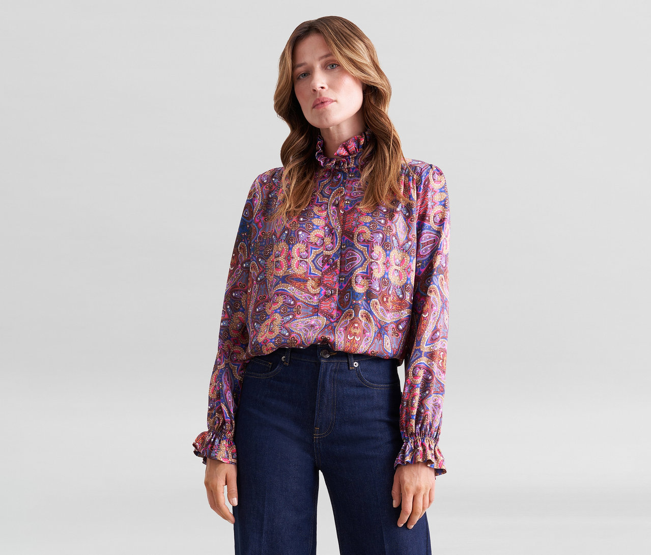 Eine Frau steht vor einem hellgrauen Hintergrund und trägt eine langärmelige Bluse mit buntem Paisleymuster, Rüschenkragen und Rüschenmanschetten sowie dunkelblaue Jeans.