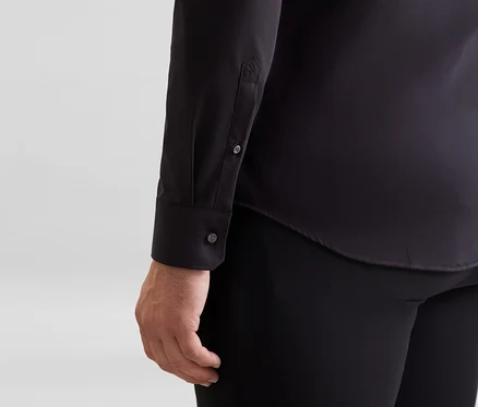 Person trägt ein schwarzes Langarmhemd mit Knöpfen an der Manschette und einer kleinen Stickerei am Ärmel, kombiniert mit einer schwarzen Hose.