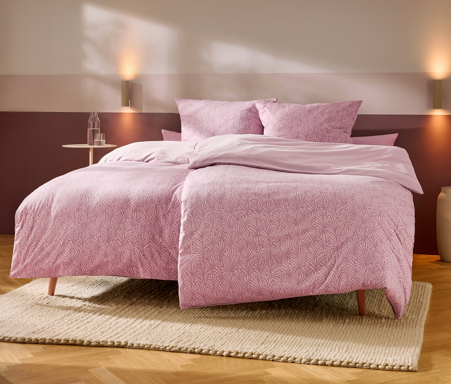 Ein Bett mit rosa gemusterten Bettbezügen und Kissen sowie unifarbenen rosa Laken steht auf einem hellen Teppich auf Holzboden. Links daneben ein kleiner runder Tisch mit Wasserflasche und Glas. Zwei Wandleuchten erhellen die zweifarbige rosa Wand hinter dem Bett, rechts steht eine hohe Vase.
