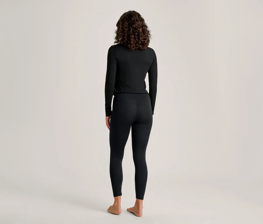 Eine Person mit dunklem, lockigem Haar steht mit dem Rücken zum Betrachter und trägt ein schwarzes Langarmshirt und schwarze Leggings. Sie ist barfuß vor einem schlichten hellen Hintergrund.