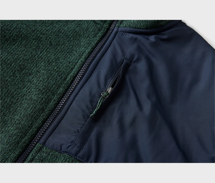 Nahaufnahme eines Kleidungsstücks mit dunkelgrünem Strickstoff links und dunkelblauem Glattstoff rechts, getrennt durch einen schwarzen Reißverschluss. Eine Reißverschlusstasche mit grünem Zugband befindet sich auf dem blauen Bereich.