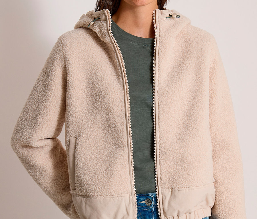 Eine lächelnde Frau trägt eine beigefarbene CECIL Jacke aus Teddyfleece mit Kapuze über einem dunkelgrünen T-Shirt und blauen Jeans.