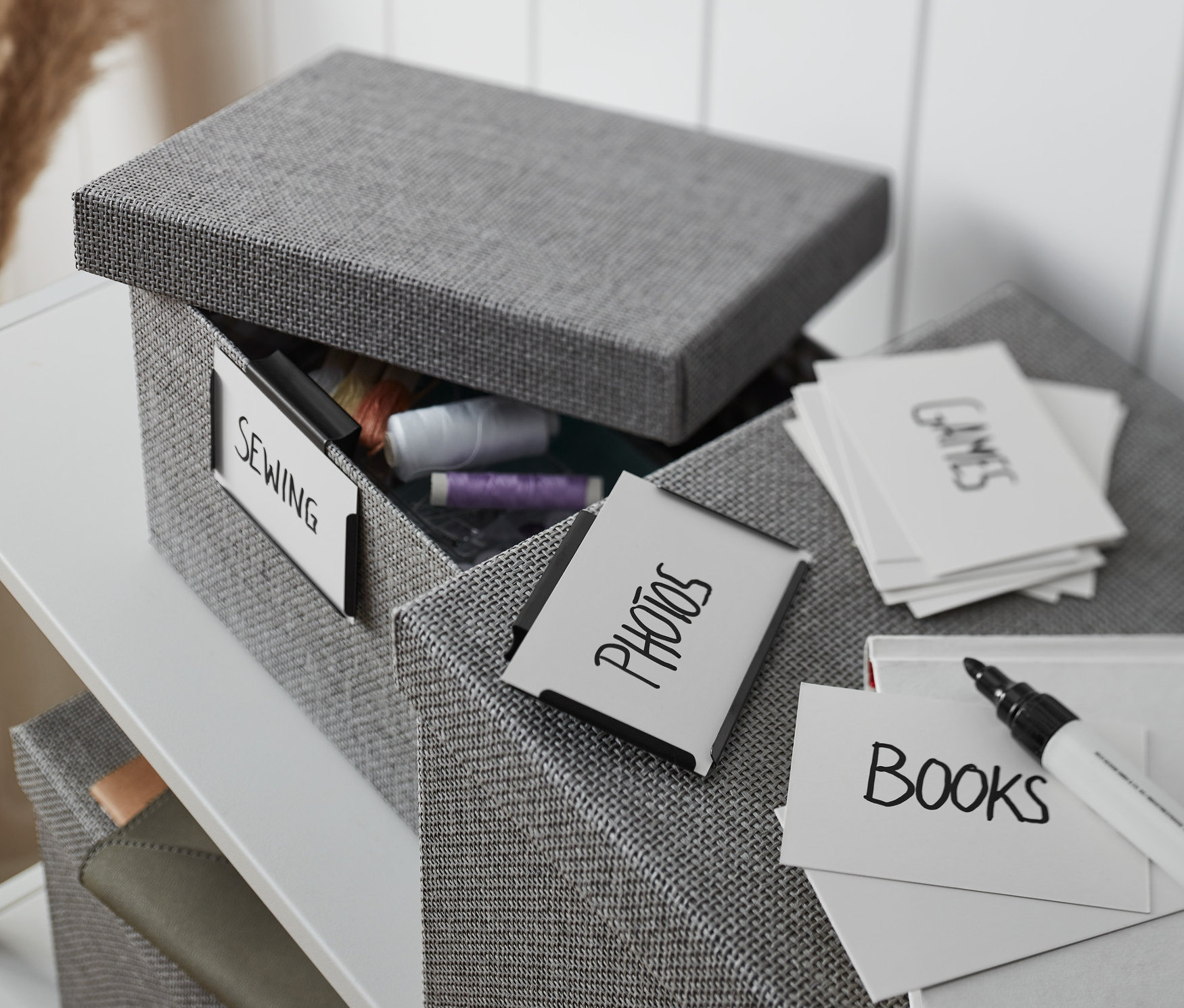 Geöffneter, grauer Stoffkasten mit 2 Metall-Clips, "Nähen"-Etikett und Fadeninhalt, daneben Kasten mit beschrifteten Karten.