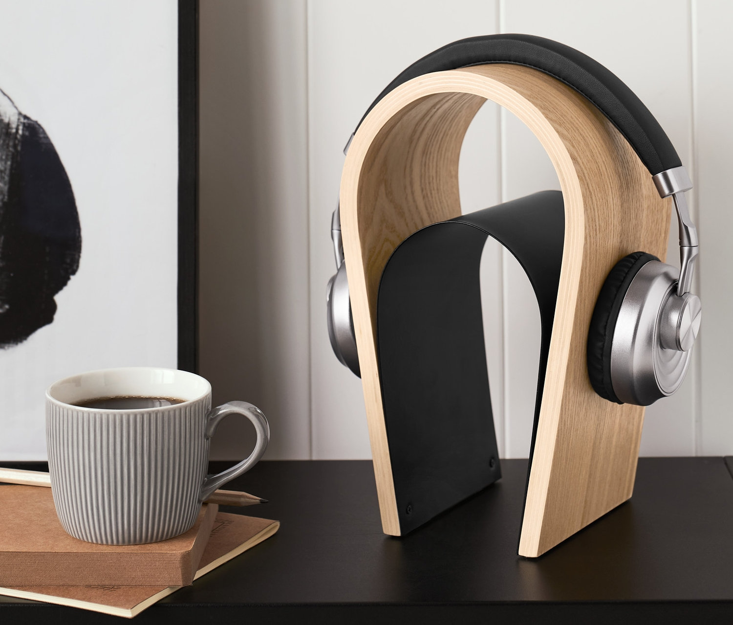 Kopfhörer auf einem Holz-Kopfhörerständer. Daneben stehen eine graue Tasse und ein Notizbuch.