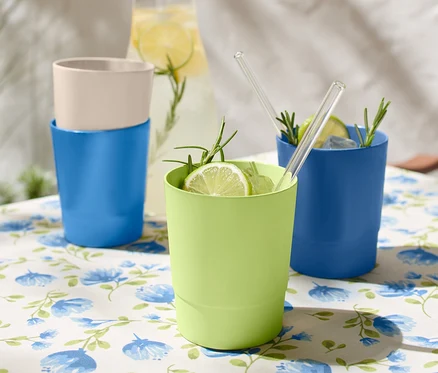 Auf einem Tisch mit blumiger Tischdecke stehen 4 Kunststoff-Trinkbecher, zwei blaue, ein grüner und ein beiger.