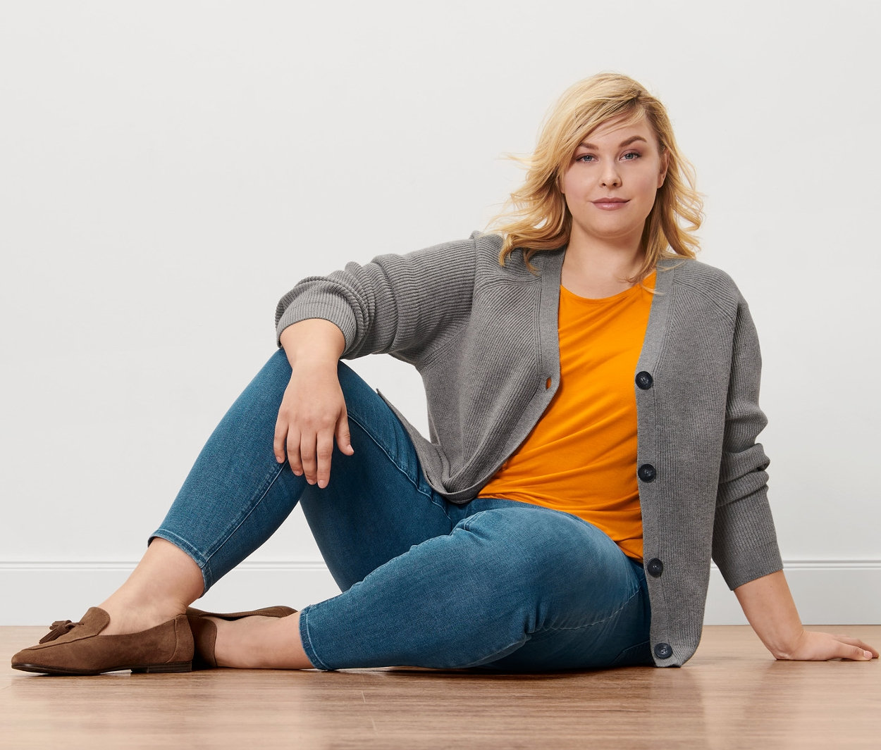 Blonde Frau sitzt auf Holzboden, trägt Treggings »Fit Nelly«, orangefarbenes Blusenshirt, Grobstrick-Cardigan und Loafer aus Veloursleder.