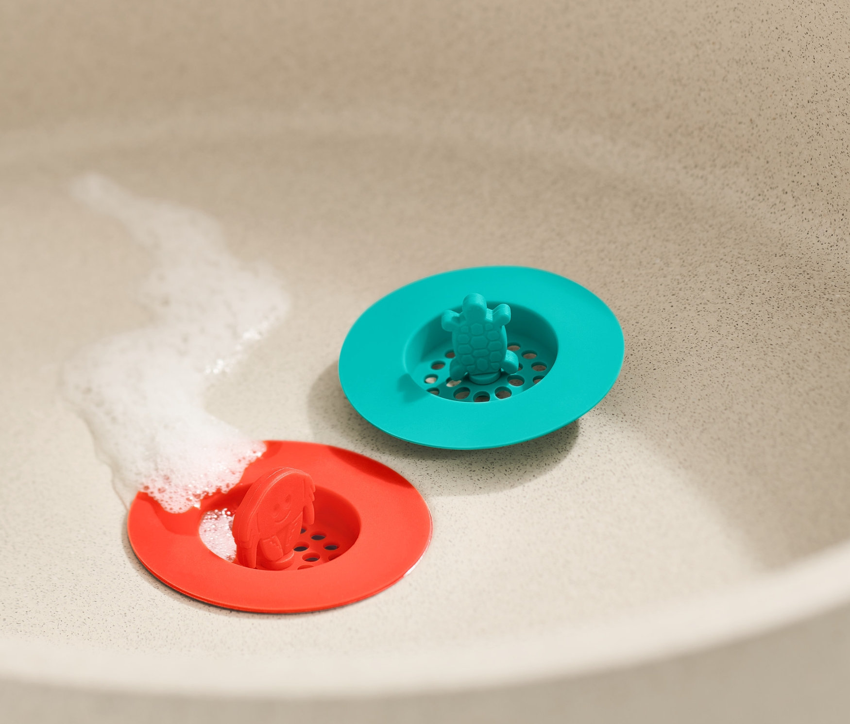 Im Waschbecken liegen zwei Abflusssiebe aus Silikon in Rot und Türkis. Unter dem roten Sieb fließt Wasser.