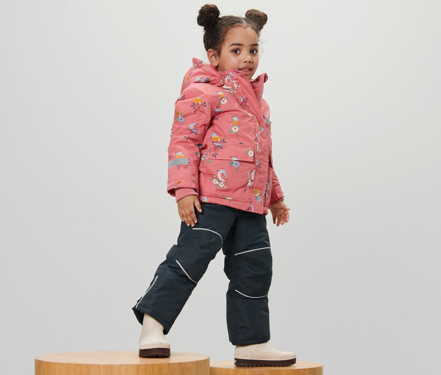 Mädchen posiert auf Holzhockern in zweiteiliger Schneekombi mit Vogel-Alloverprint.