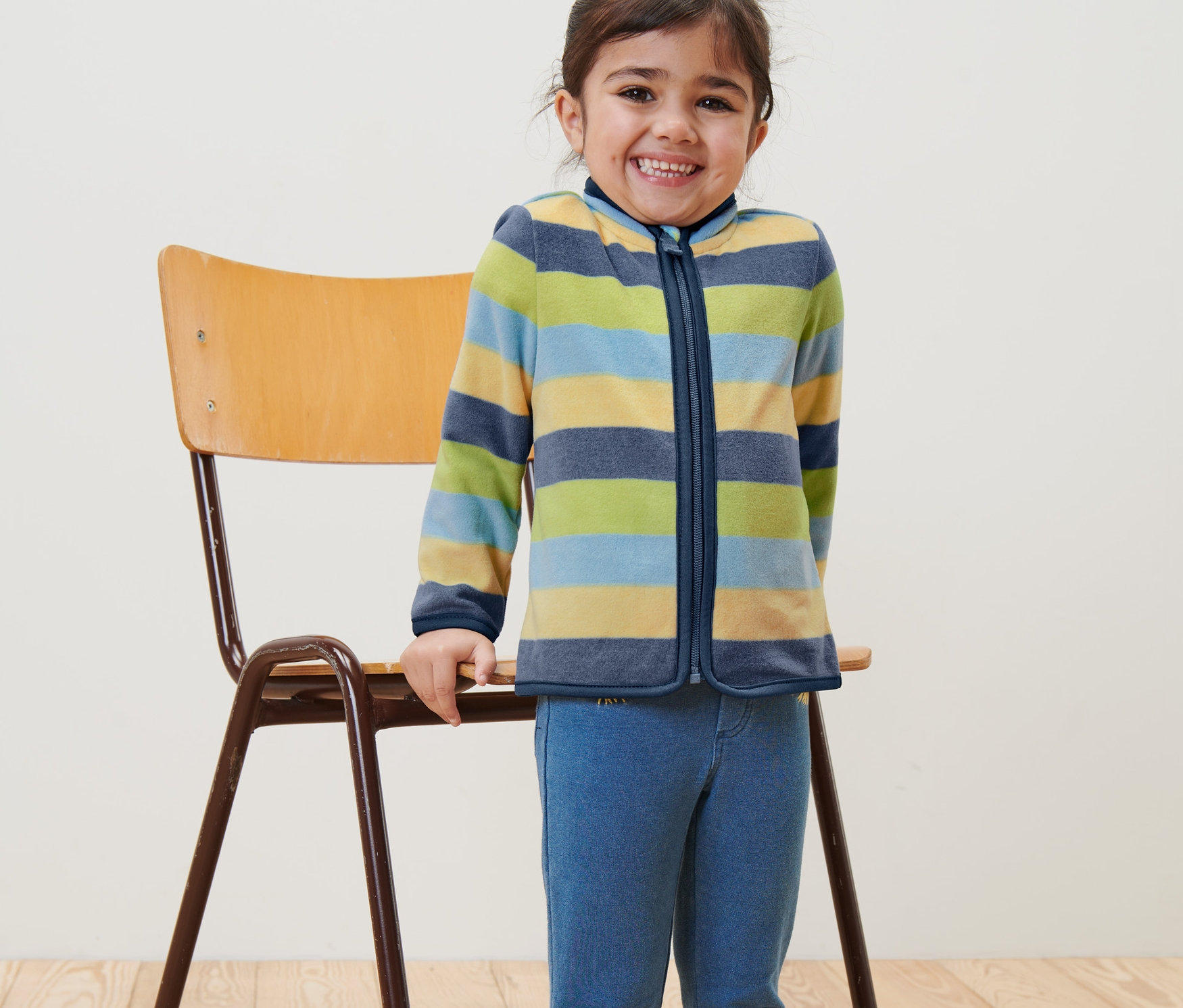 Ein lächelndes Mädchen steht neben einem Stuhl und trägt eine Kinder-Fleecejacke mit Streifen.