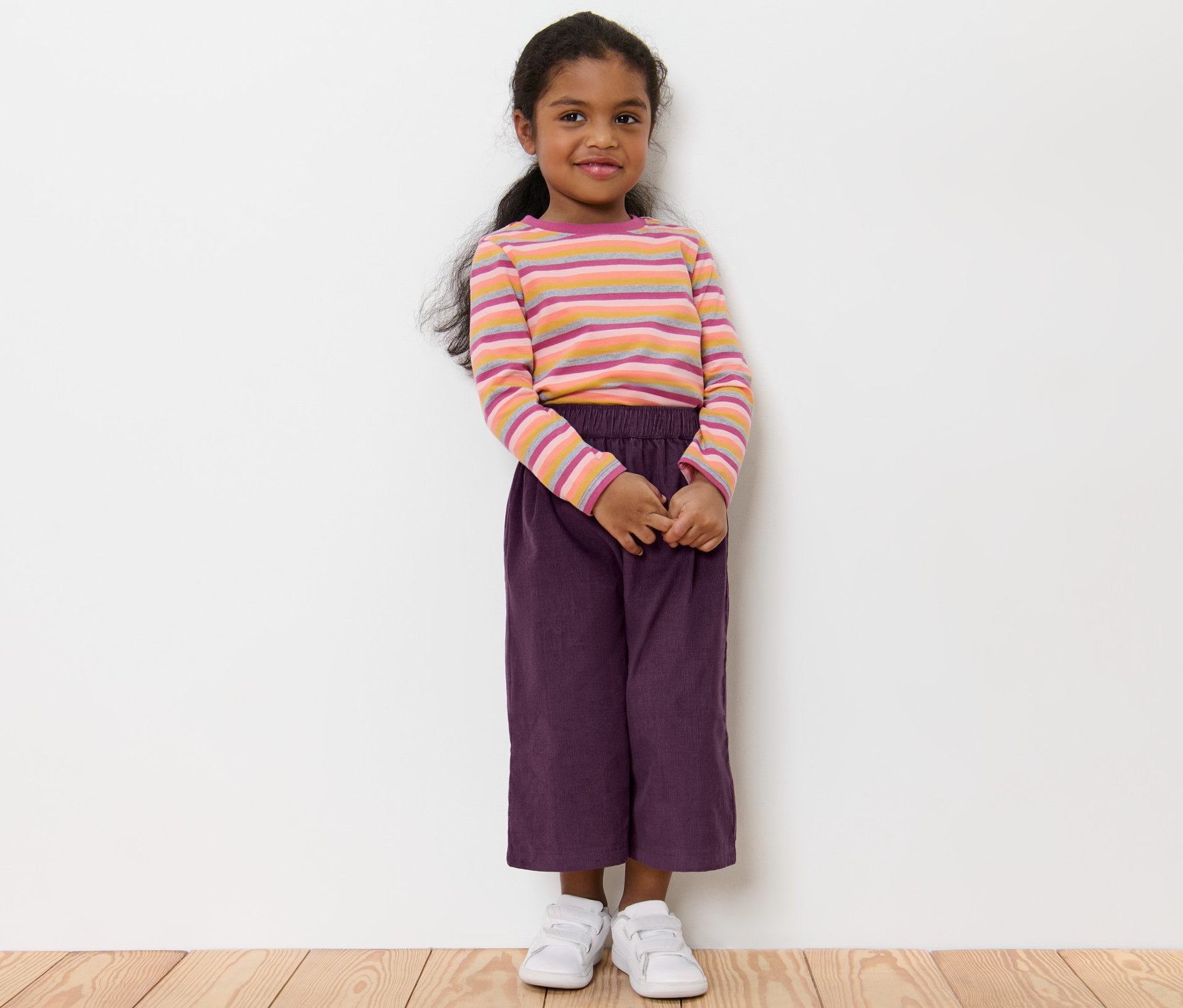 Ein Mädchen steht und trägt eine Kinder-Culotte, ein gestreiftes T-Shirt und weiße Schuhe.